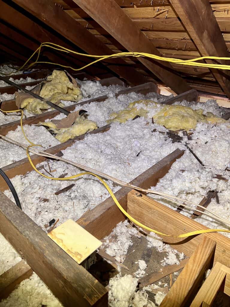 Attic space with exposed wooden rafters, yellow electrical cables, and fluffy insulation on the floor.
