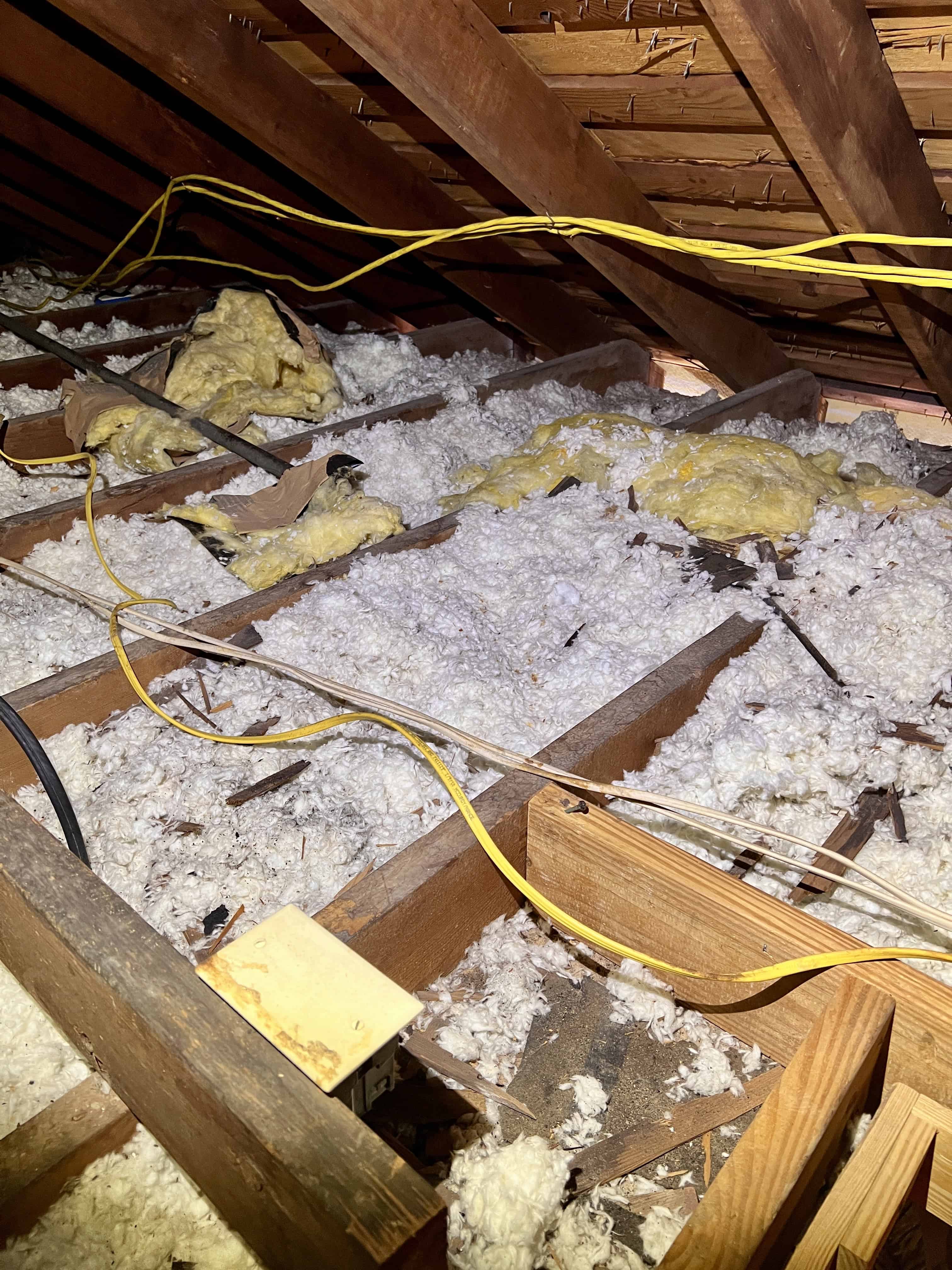 Attic space with exposed wooden rafters, yellow electrical cables, and fluffy insulation on the floor.
